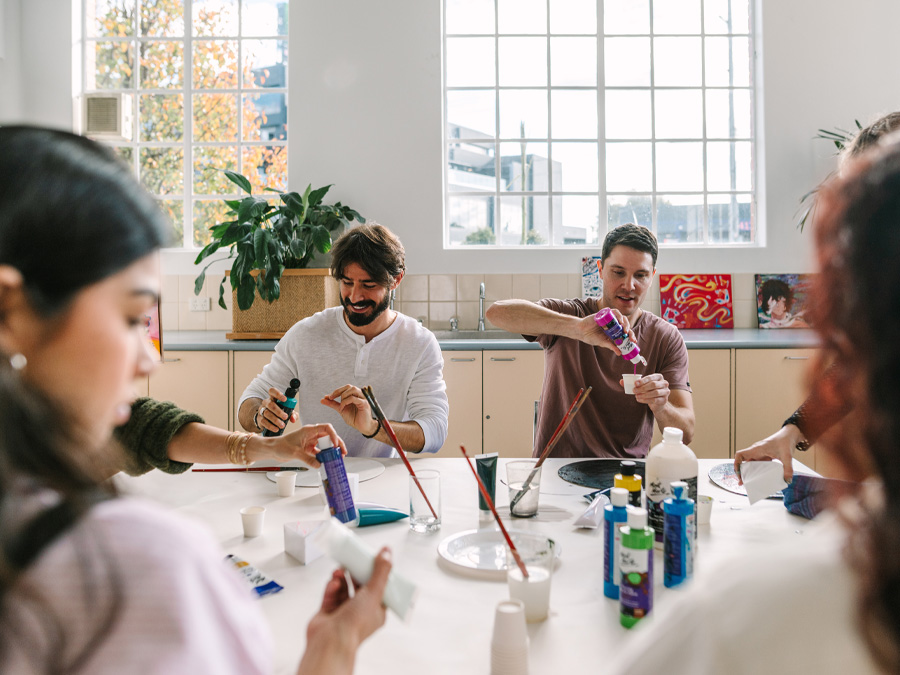Two people sit around a table in the Kingston Arts Centre, participating in a creative art-making workshop. The workshop instructor is standing in the centre of the photo, demonstrating a paint technique. Paint and paint brushes are on the table.