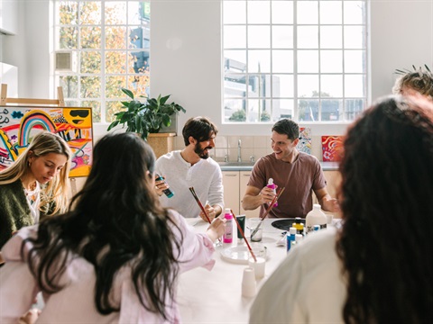 Five people sitting around a table in the Kingston Arts Centre, participating in a creative art-making workshop. The workshop instructor is partially obscured in the background. Paint and paint brushes are on the table.
