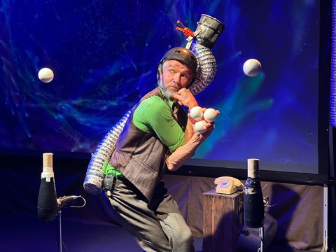 Jens Altheimer wearing a green shirt and a bike helmet with junk attached to it, juggling 5 balls between hair-dryer contraptions.