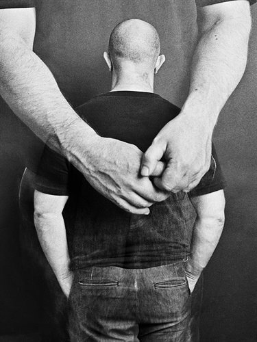 A black and white manipulated photograph of a person wearing a black shirt and jeans facing away from the camera. Large, clasped arms are superimposed across their back, as if in an embrace.
