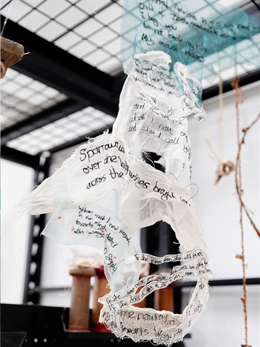 a photograph of a delicate fabric artwork with stitched text hanging from a metal wire shelf