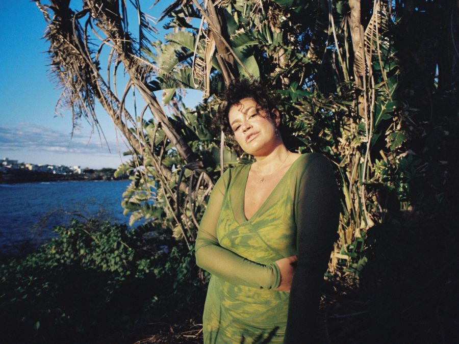 A dark haired female wearing a long green dress standing against tree with body of water in the background