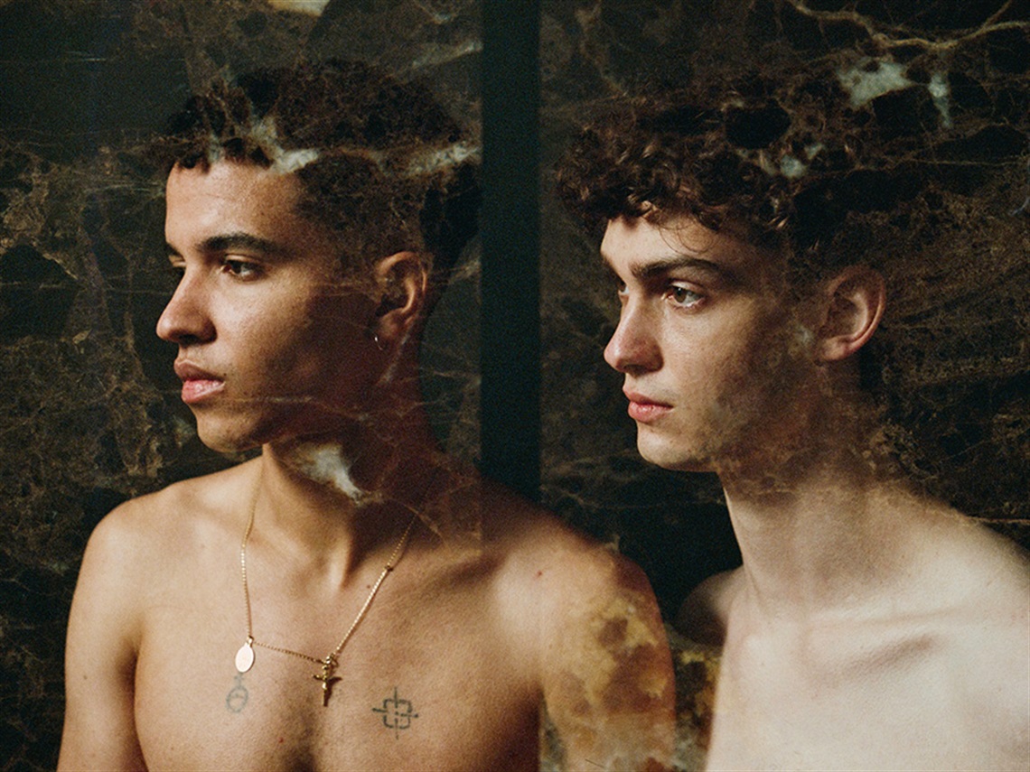 a close up photo of two young men with bare chests looking to the left. There is another overlay of abstract imagery over the top. 