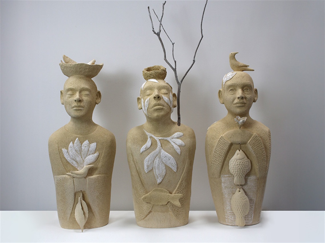 Three ceramic bust sculptures by Michelle Merrifield standing side by side. The figures have imagery of birds, fish, baskets, and plants carved into, or on their bodies.
