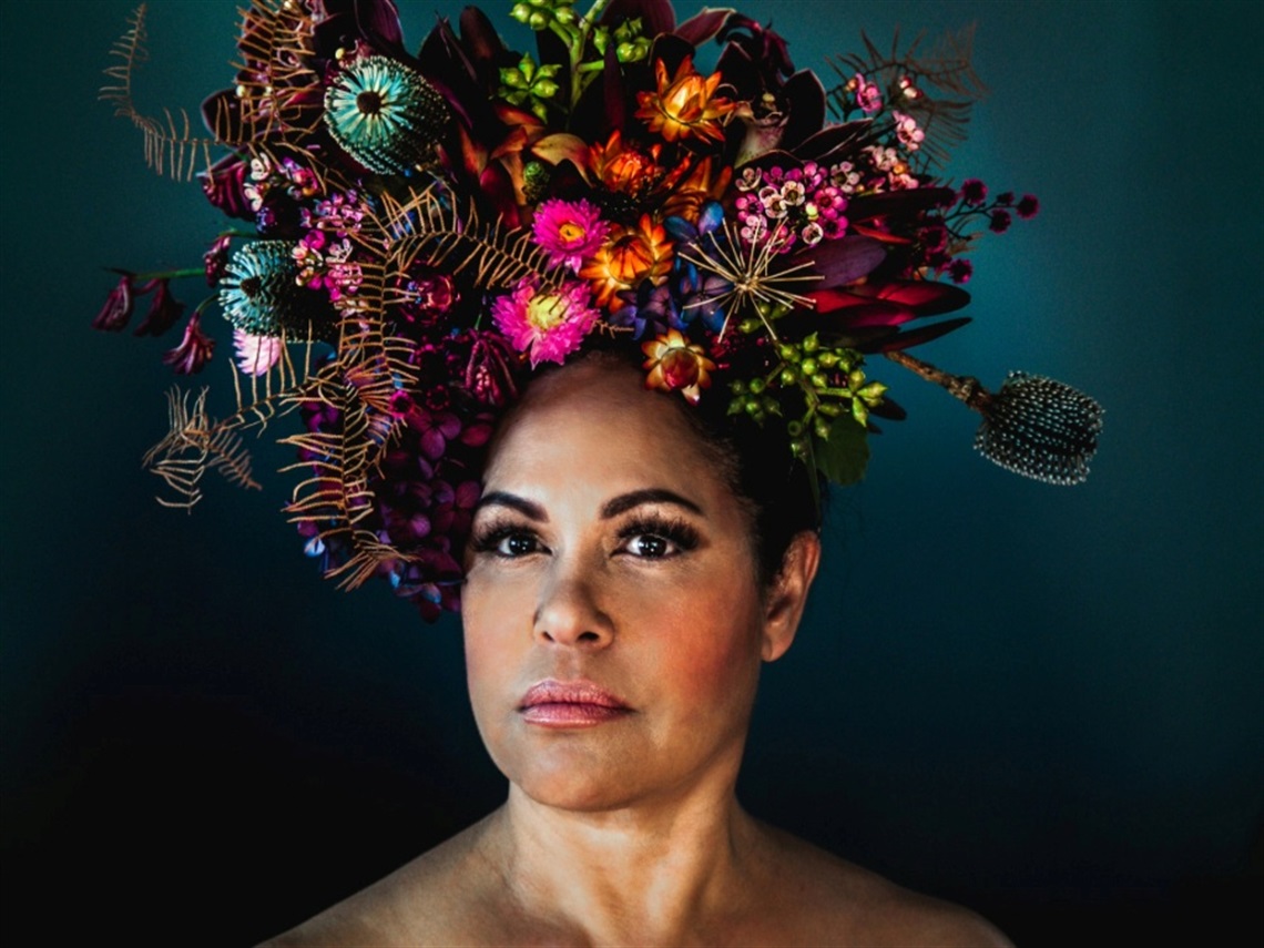 A woman is in front of a dark teal background and wears various native Australian flowers in purple and blue around her head like a crown.