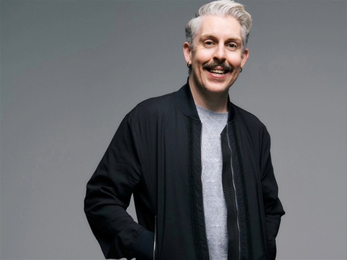 A man smiling wears a black jacket and grey shirt.