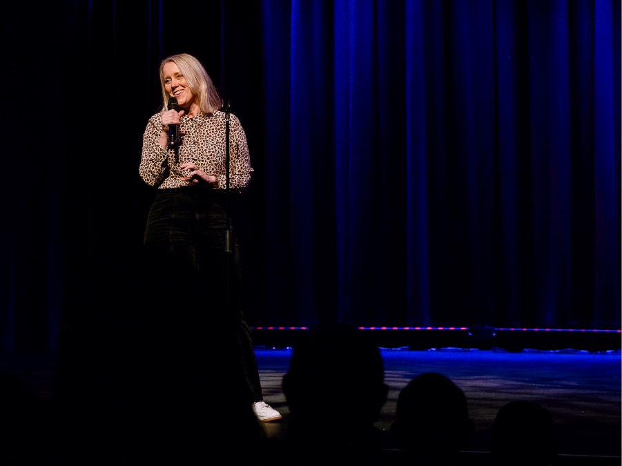 A woman stands on a theatre stage with blue lights and an audience watching.
