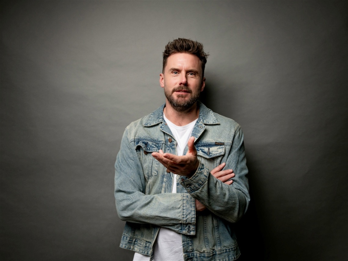 Comedian Dave Thornton standing in front of a grey background wearing a white t-shirt and denim jacket.