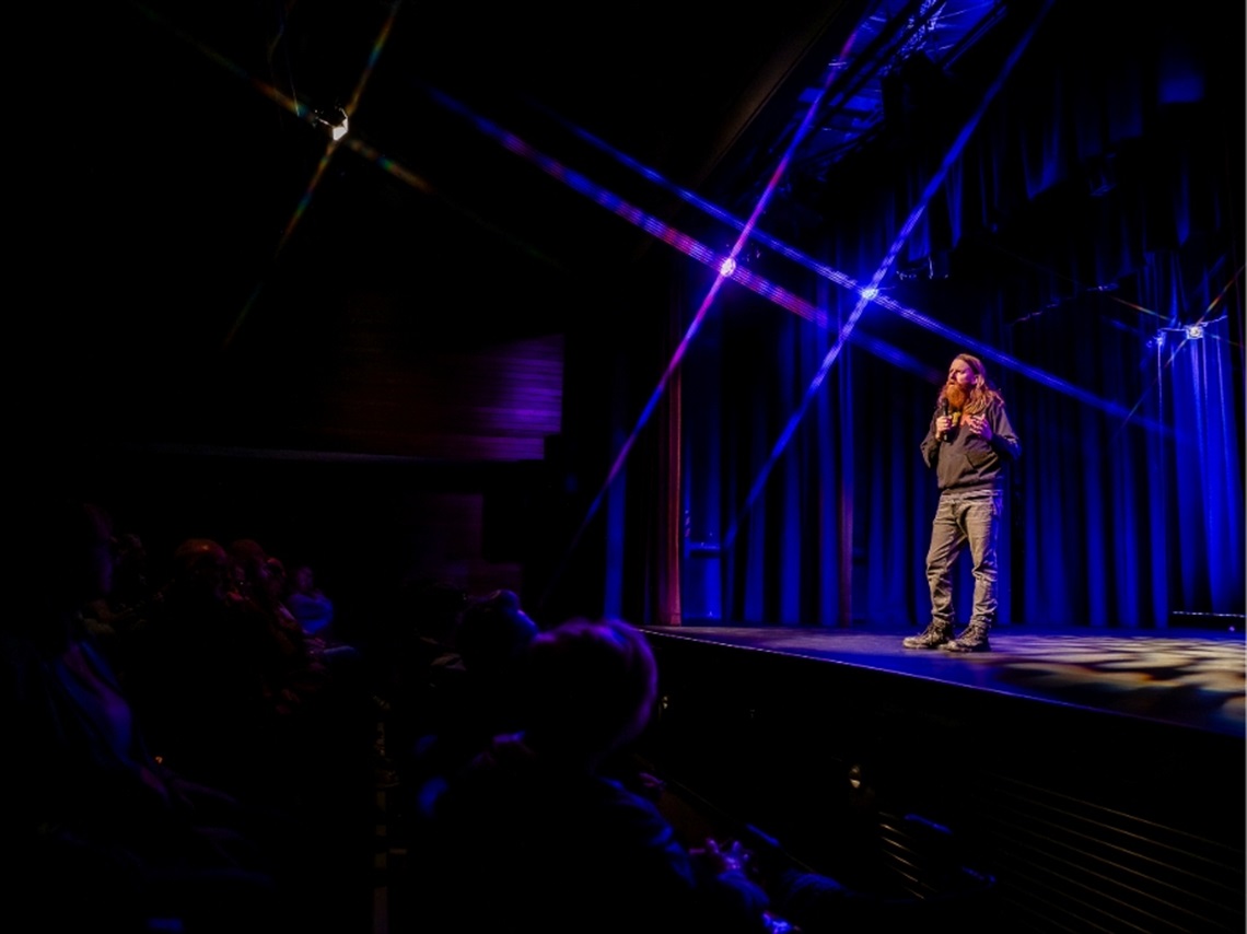 A man standing on a stage with blue stage lights and an audience watching.