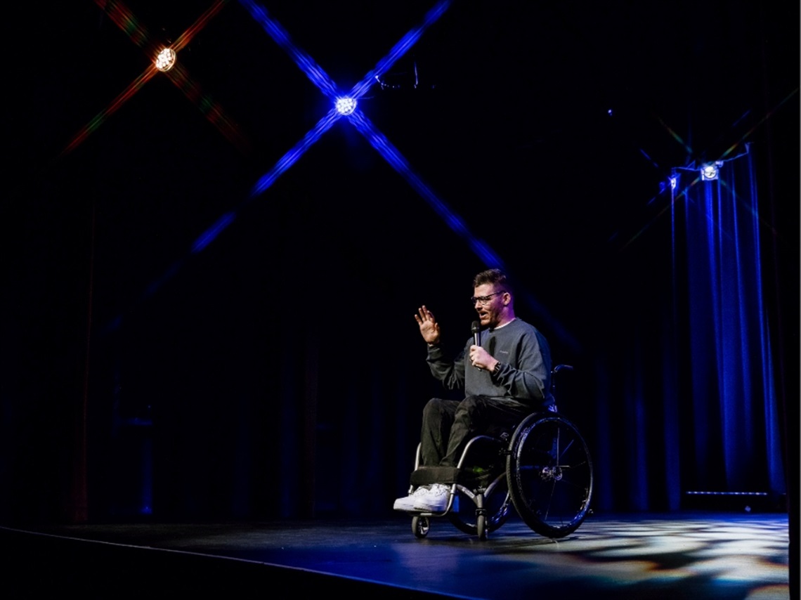 Man in a wheelchair on a stage holding a microphone with blue stage lights behind him.