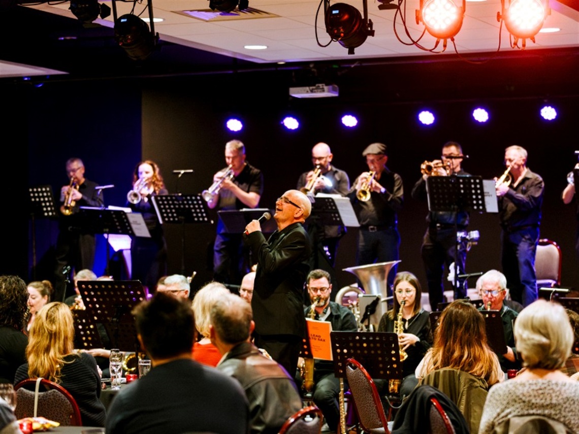 A man sings with a microphone in front of a jazz band while a seated audience watches.
