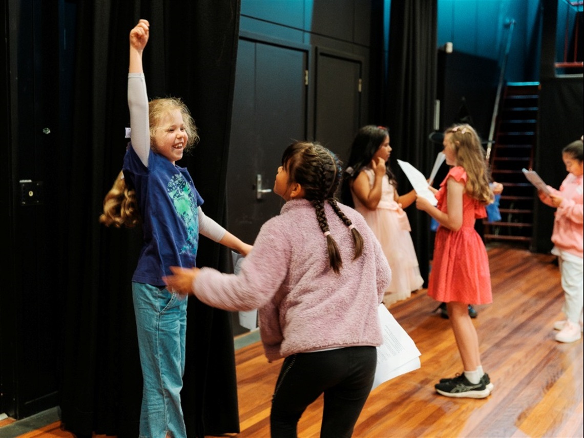 Children dance and read a script in a theatre.