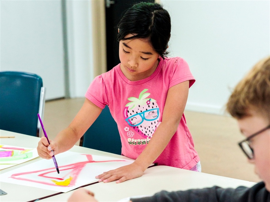 A young girl in a pink tshirt using a paintbrush to paint a pink pattern on paper.