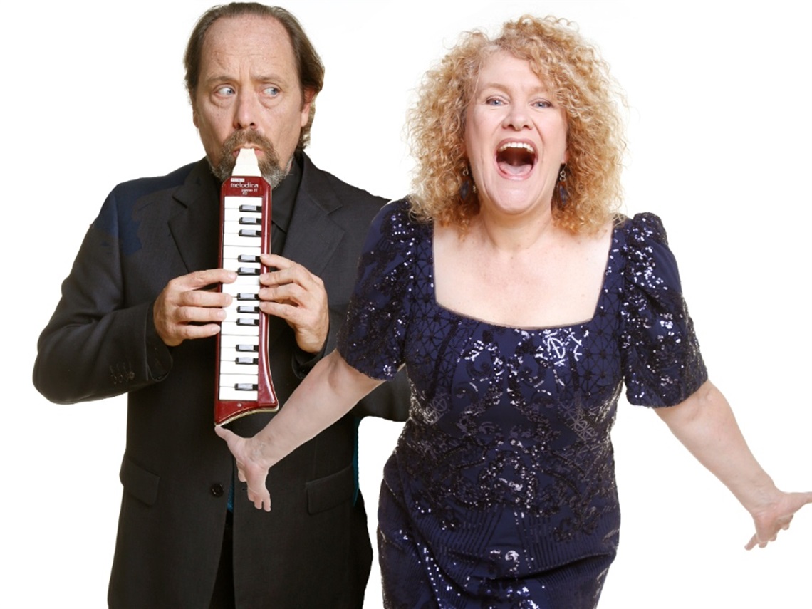 A man wearing a black suit holds an instrument and a woman in a blue glittery dress faces the front with her arms outstretched