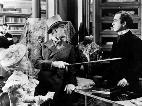 A man and woman sit in a tailor shop and the man has his walking stick pointed at the shop assistant. 