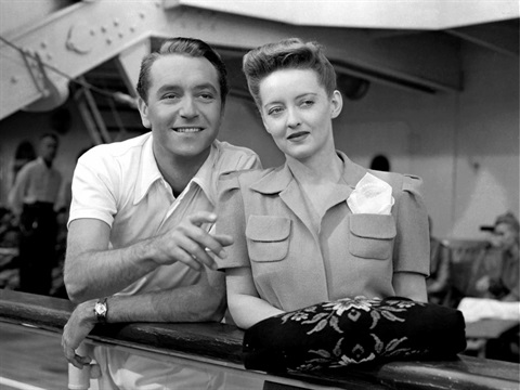 A man and woman stand at the edge of a ship railing smiling