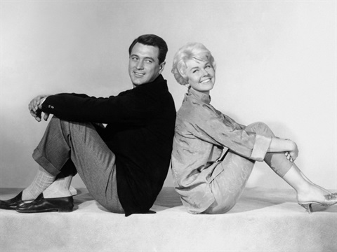 A man and woman sit on the floor with their back against each other smiling