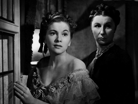 Two women stand in a doorway. One woman is taller and both women are staring to the right