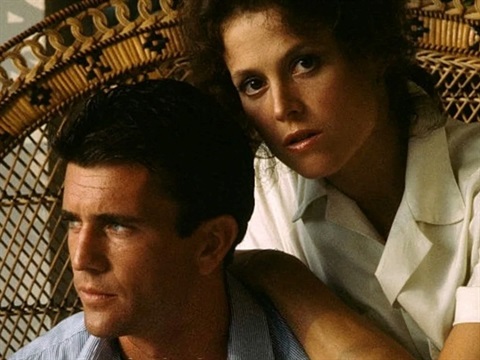 A man and woman sit on a rattan chair. The man is looking to his right and the woman stares forward with a serious expression