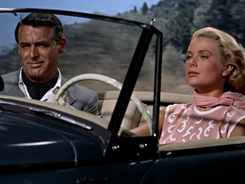 A woman driving a car with a man next to her. The woman is wearing a pink scarf and the man is wearing a grey suit
