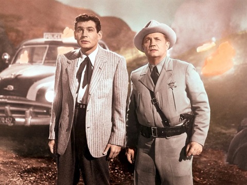 Two men stand with shocked faces with a taxi behind them and hills on fire