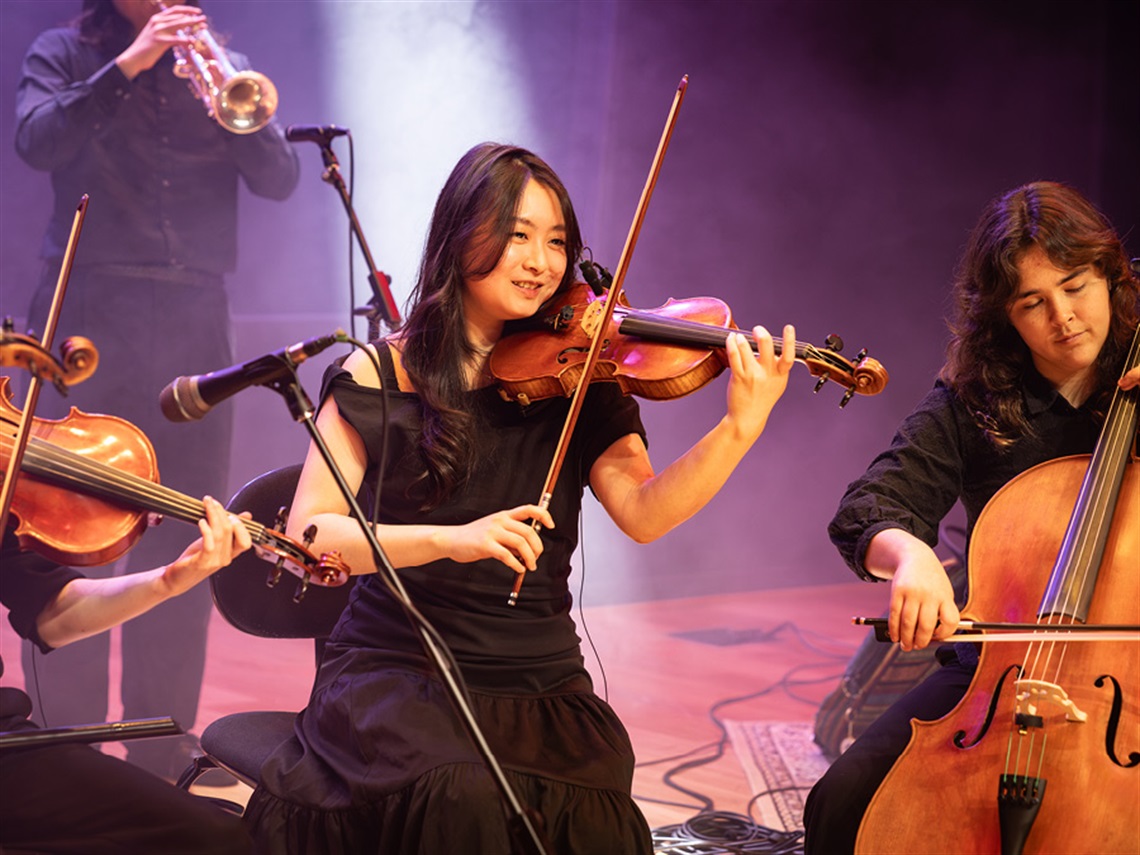 A violinist smiling and wearing all black while playing on stage, surrounded by other string and brass musicians.