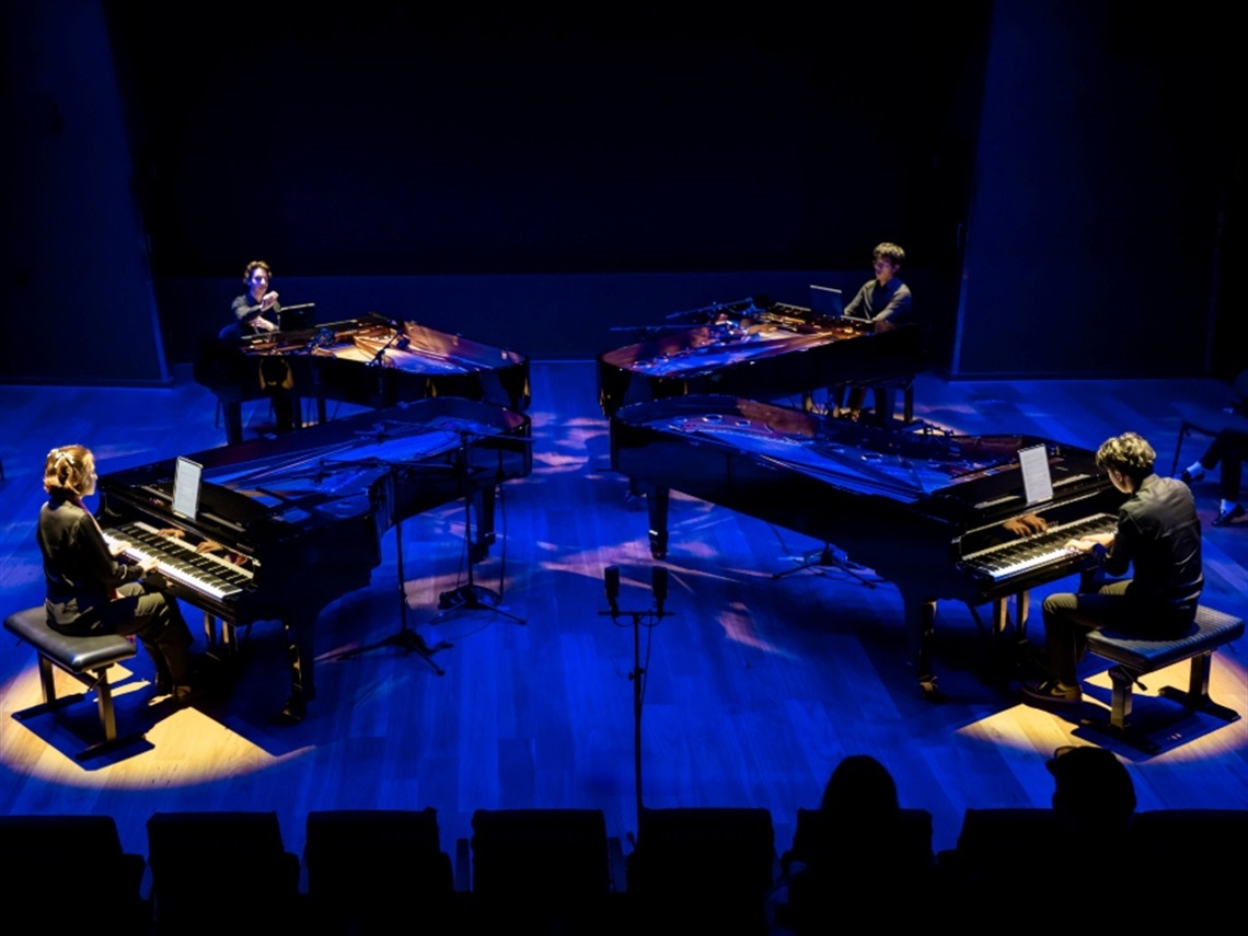 Four pianists perform on grand pianos arranged in a circle under blue stage lighting.