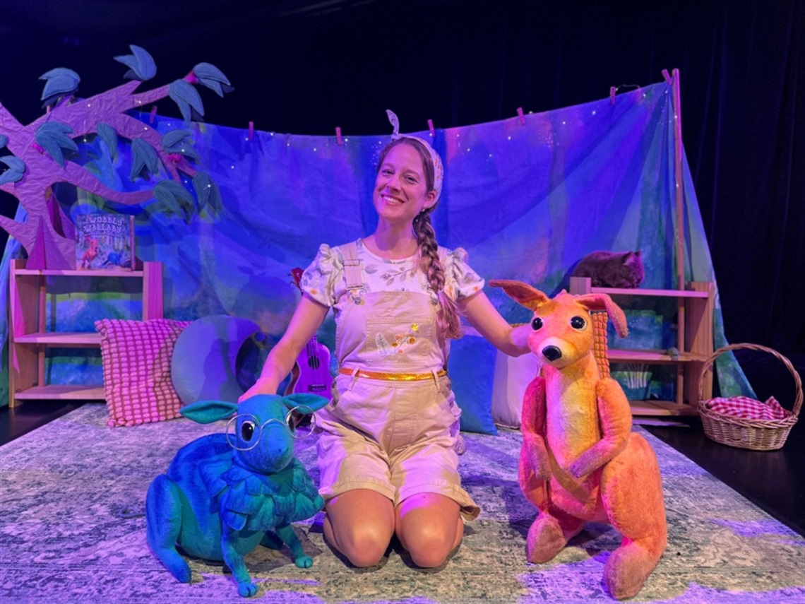 A woman sits in between a blue bandicoot puppet and an orange wallaby puppet on a rug.