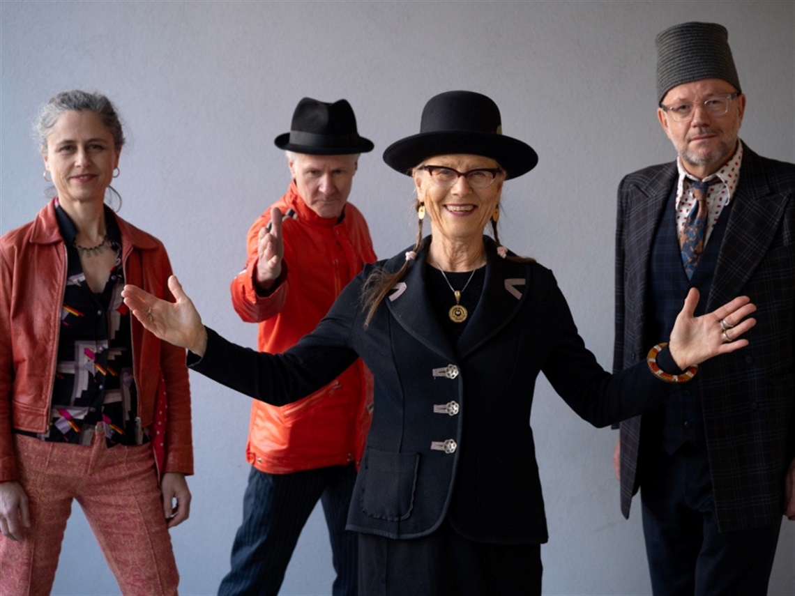Four band members stand next to each other in various poses. Three band members wear hats.