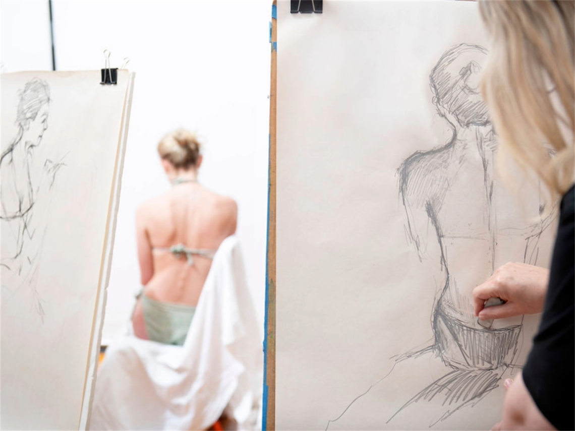 Artist drawing a model in a life drawing workshop, using a pencil on paper attached to an easel
