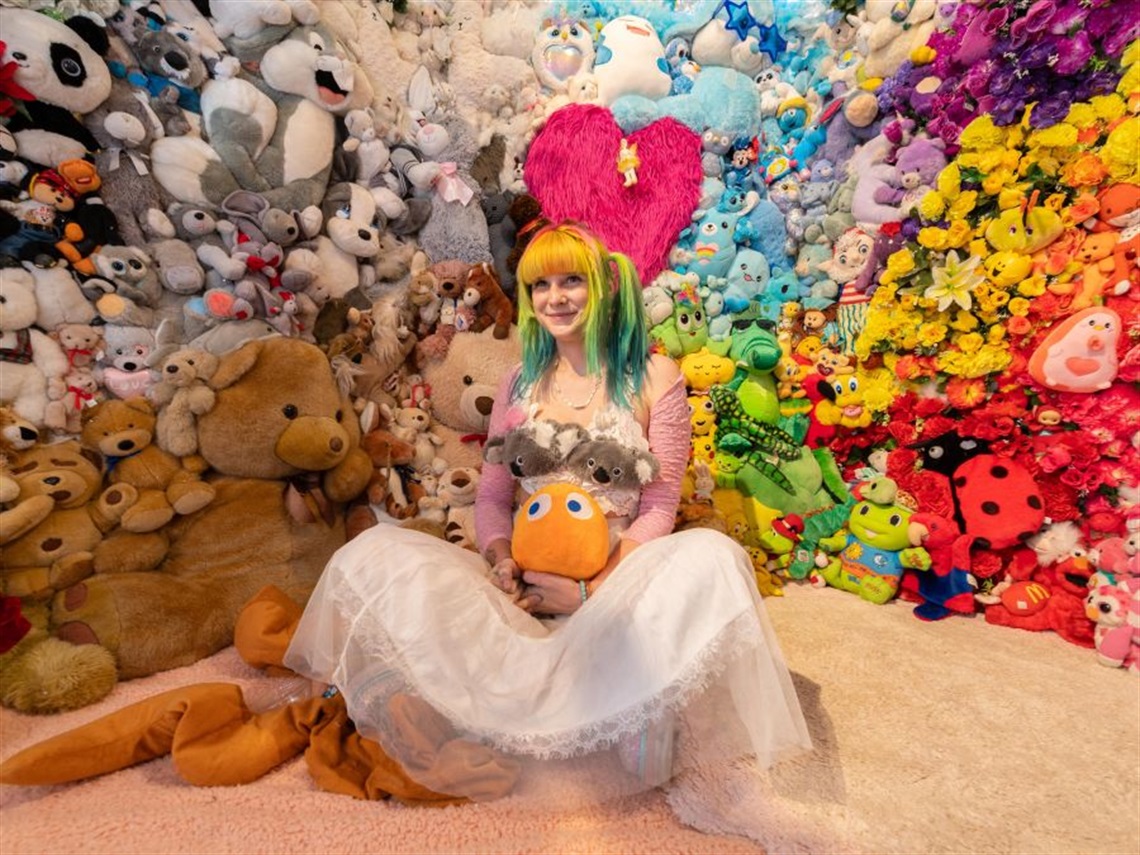 Female with multicoloured hair wearing a white skirt, holding several stuffed toys, seated against a backdrop of multiple colourful stuffed toys
