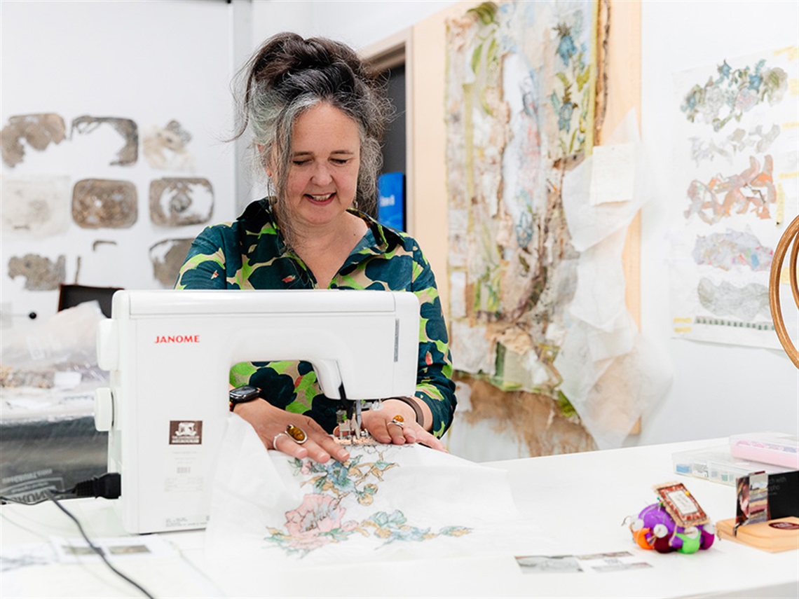 Mid-view of a woman wearing a black and green shirt, standing and using a white sewing machine in an artist's studio