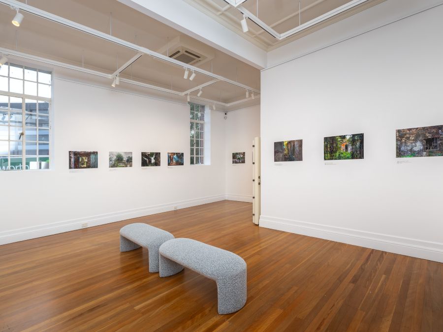 A gallery space with a horizontal row of colour photographic images on white walls with two light gray fabric benches on wooden floor