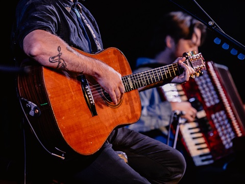 A close up of an artist strumming a guitar. Another performer holds a piano accordion in the background.