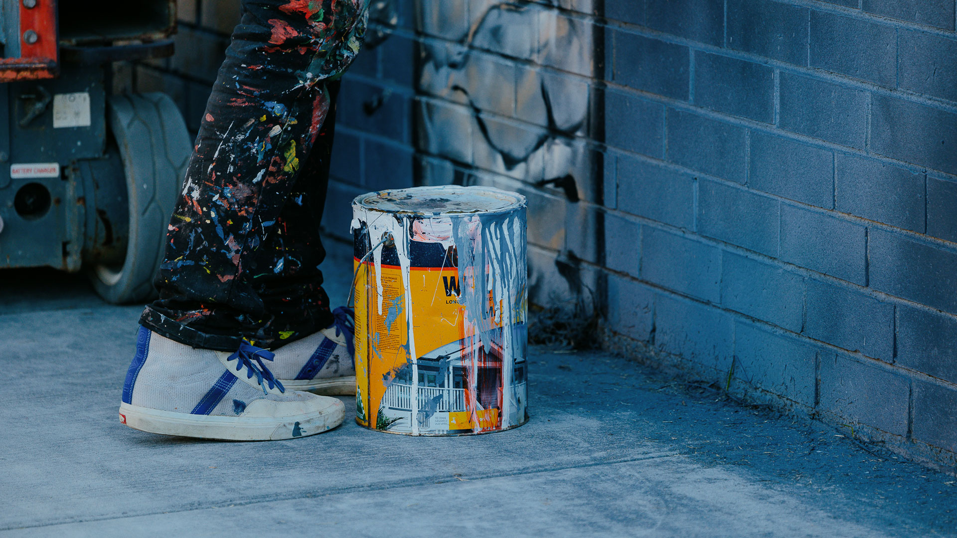 Person's feet standing near a brick wall. A paint bucket is between the feet and the wall, with paint dripping down it. 