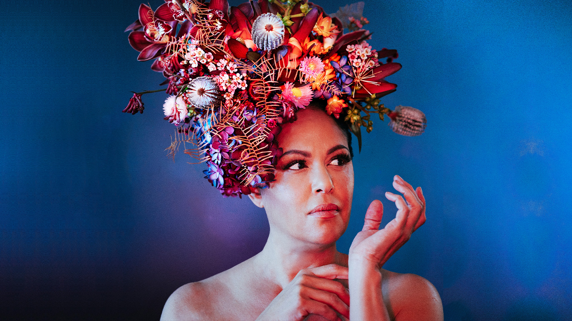Christine Anu posing against a blue background, she wears a headdress of native flowers.