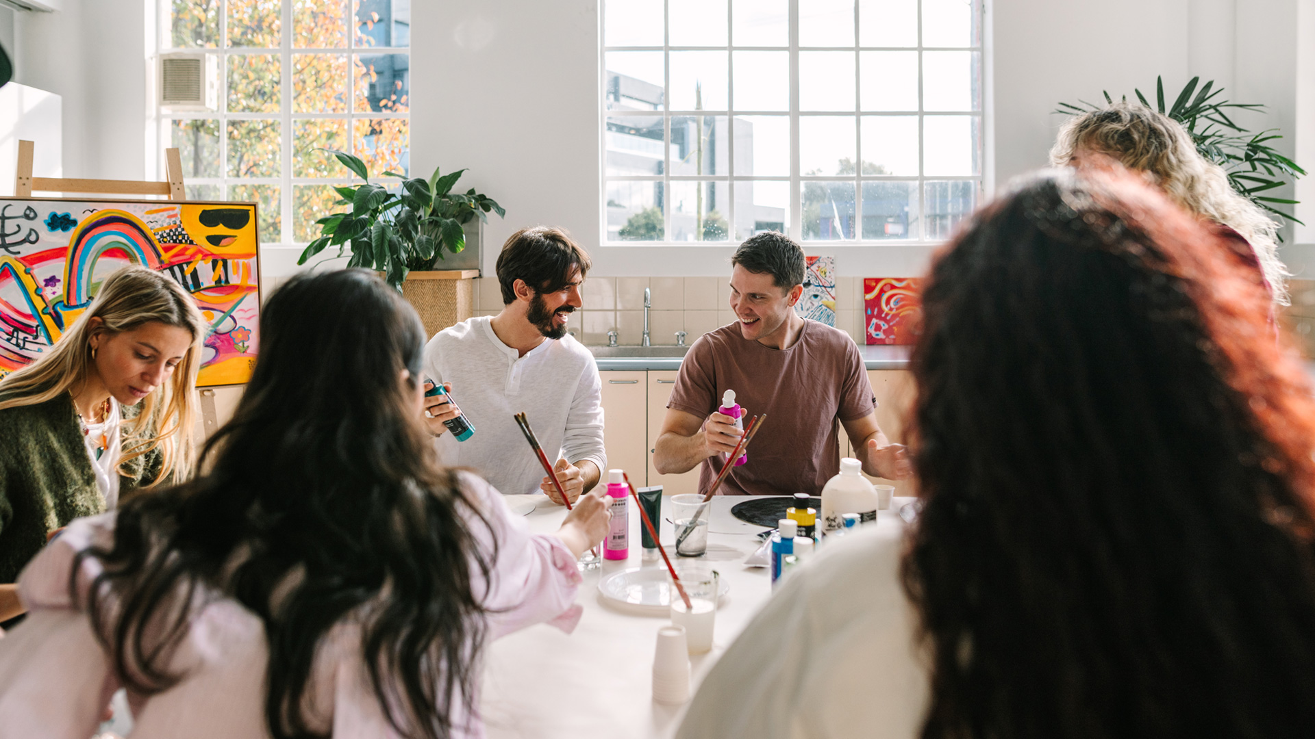Five people sitting around a table in the Kingston Arts Centre, participating in a creative art-making workshop. The workshop instructor is partially obscured in the background. Paint and paint brushes are on the table.