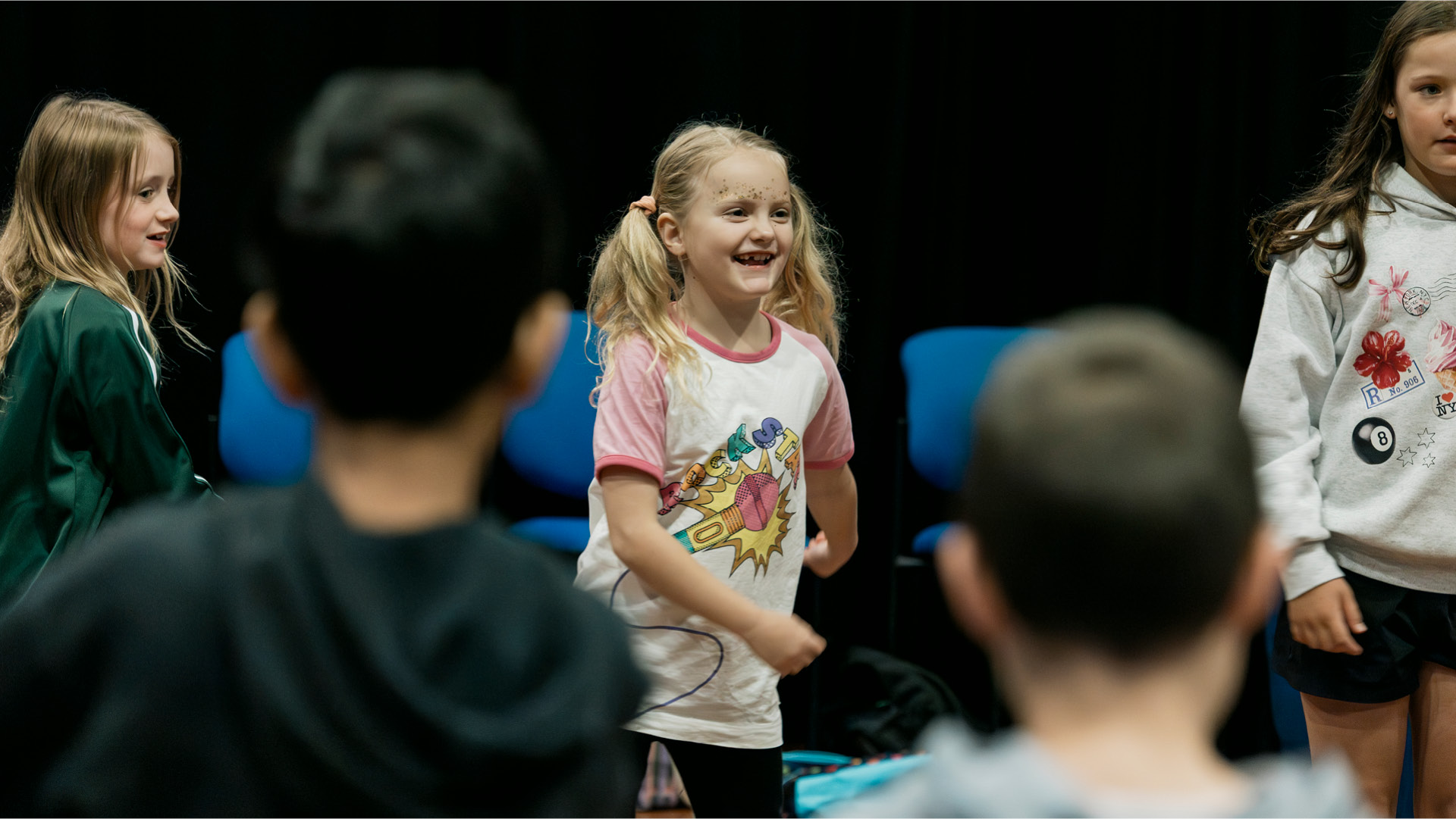 A child with pigtails and glitter face-paint smiling and dancing amongst other children in a performing arts workshop.