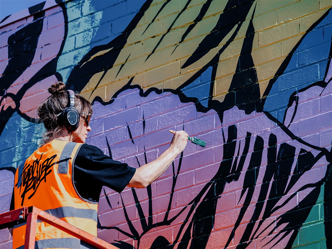 Artist George Rose painting her mural for Wall to Wall Festival 2024. Using a brush to paint florals on a brick wall using pinks, purples, blues yellows and black lineart.