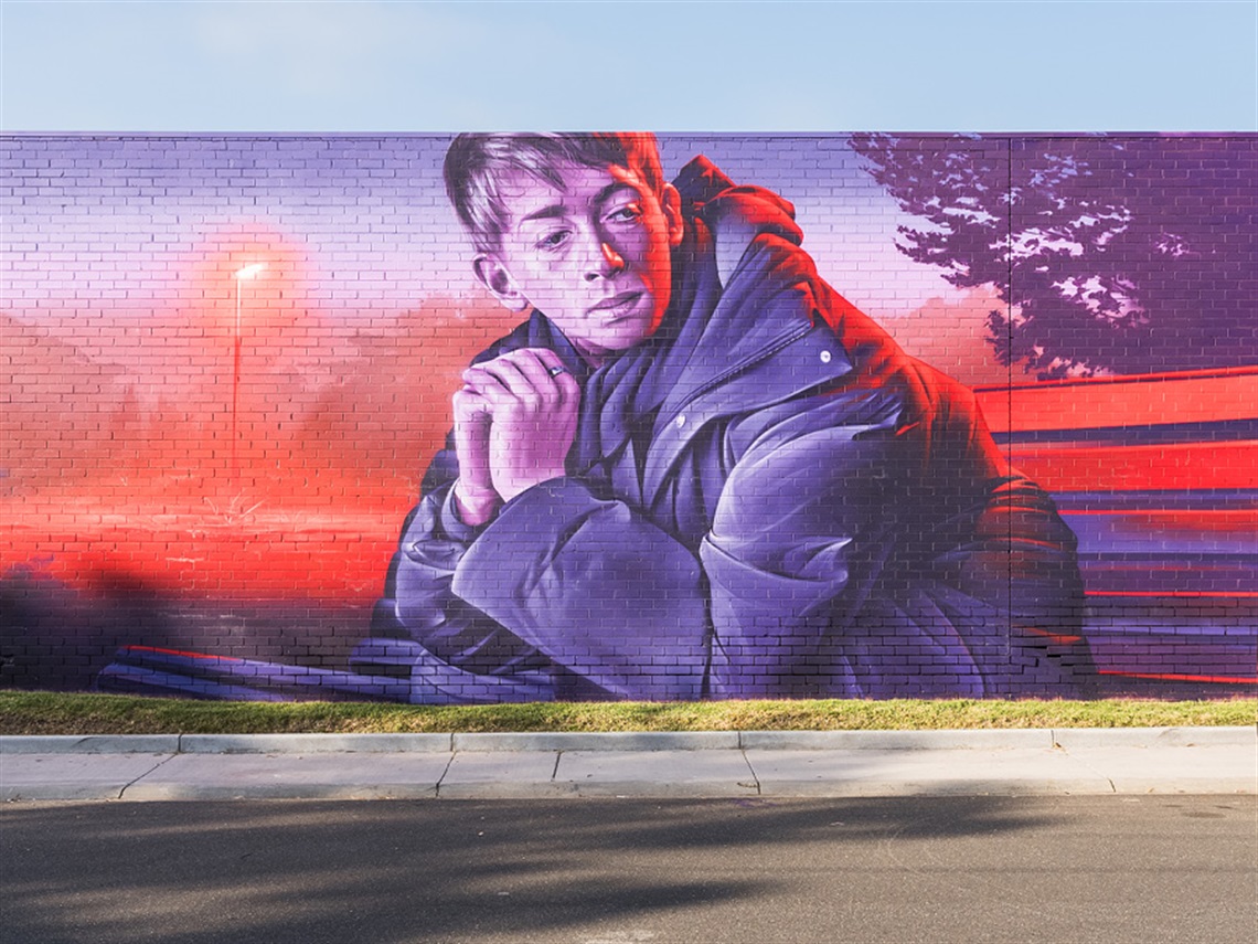 Artist Smug's mural for Wall to Wall Festival 2024. A young man sits on a park bench looking pensively over his shoulder. The mural has a sunset theme, bathed in purples and oranges.
