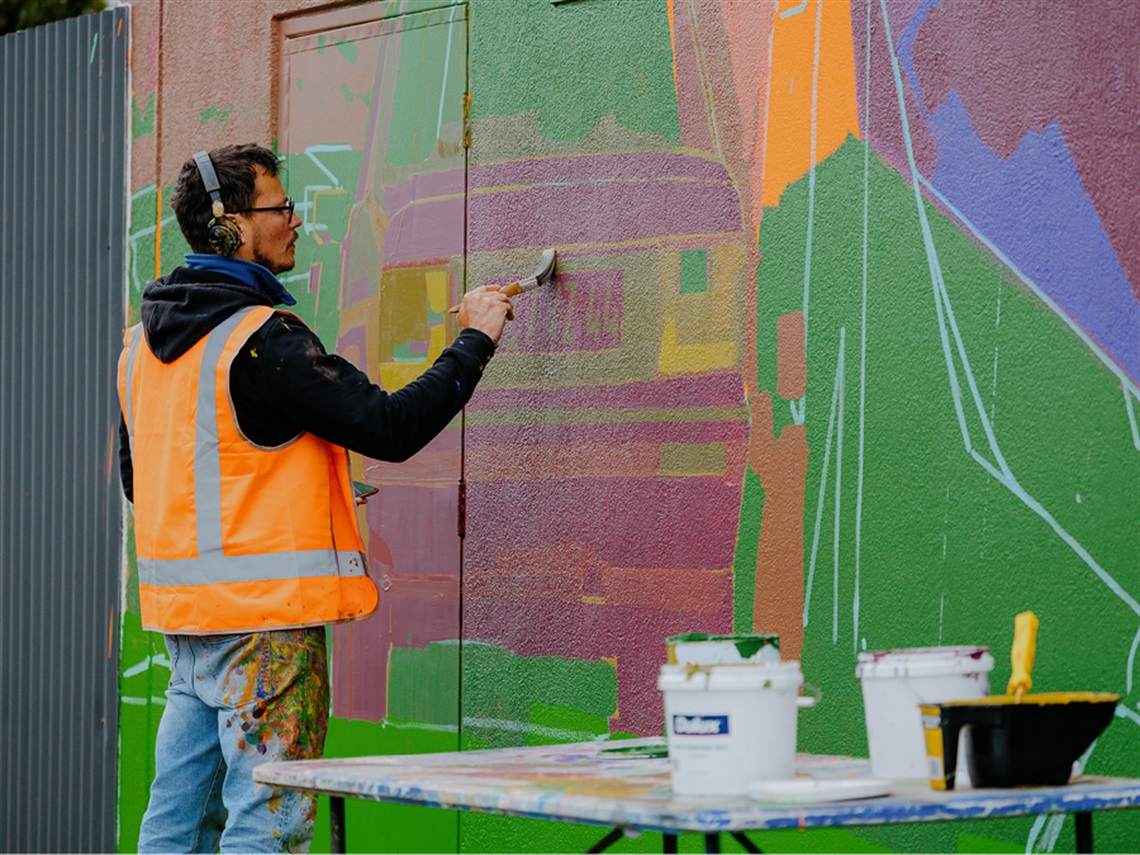 Artist Zoer's painting his mural for Wall to Wall Festival 2024. The artist makes green brush strokes over an unfinished painting of a car.