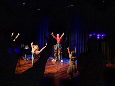 Colourfully dressed children's performer stands on the Black Box Theatre stage under warm lighting with purple accents, arms held high with two thumbs up. Children in the audience mimick.