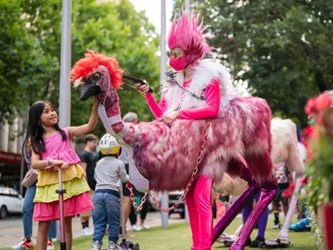 A roving performer dressed as a bright pink 'punk emu' letting a child pet the emu's neck.