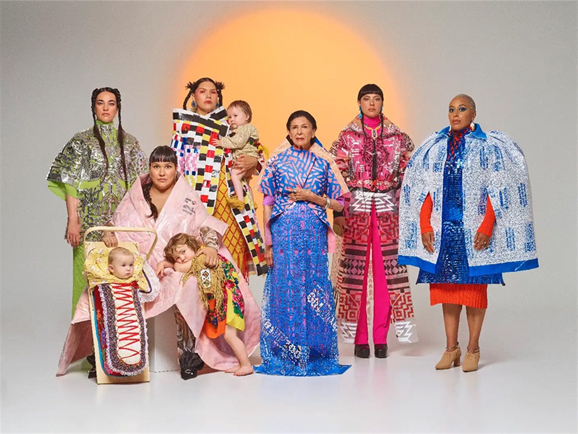 A photo of six women with three young children dressed in elaborate and colourful clothing, starting straight at the camera without smiling, against a grey and yellow background