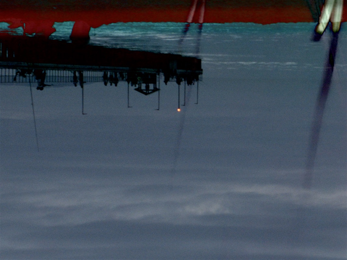 An upside-down video still of a pier to the left of image, against a dark water and night sky, with a red streak along top of image