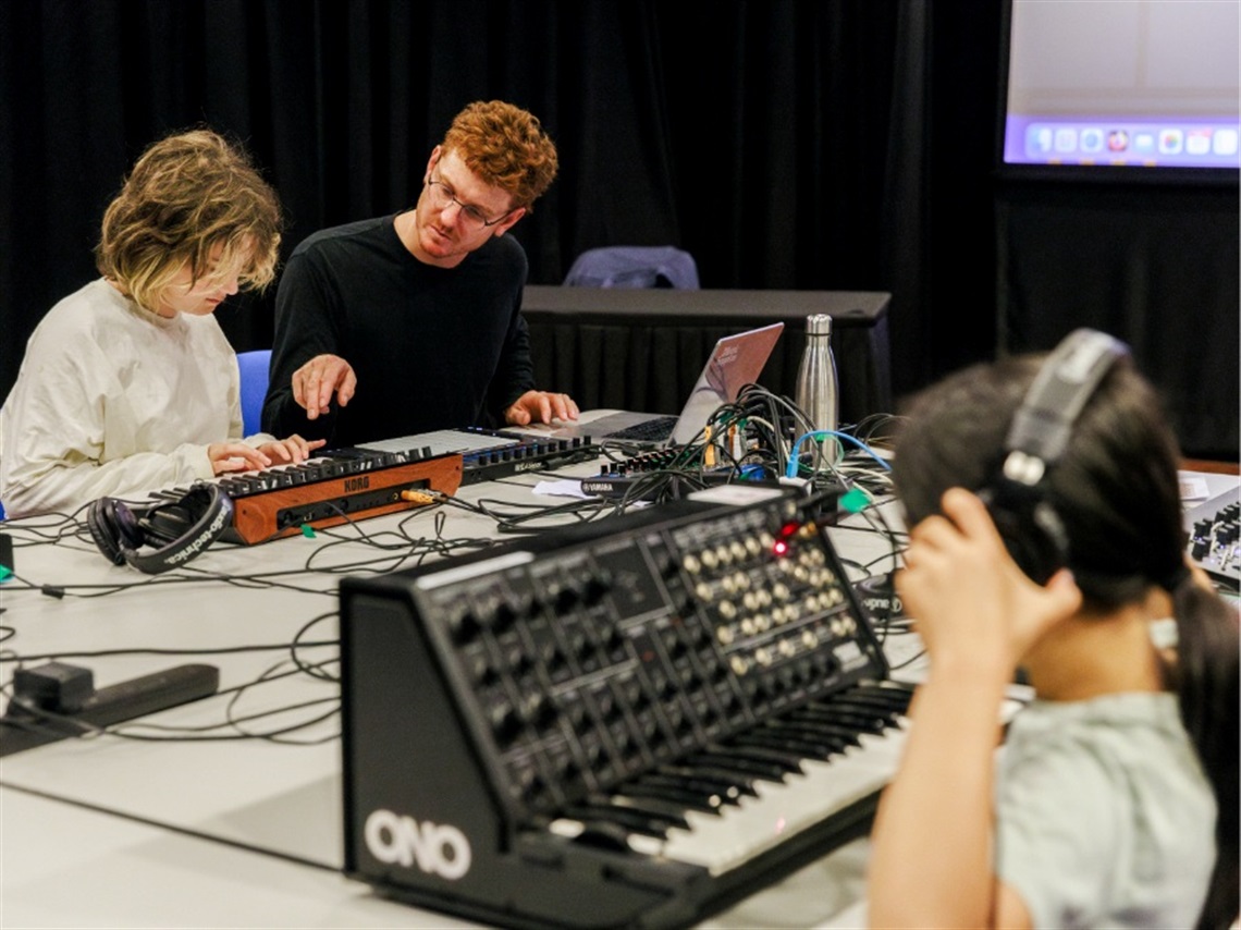 Children sit at a table with synthesiser music equipment and headphones while a facilitator teaches.