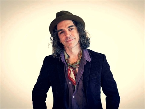Nathan Cavaleri in a hat and dark jacket posing against a plain background with a scarf at the neck.