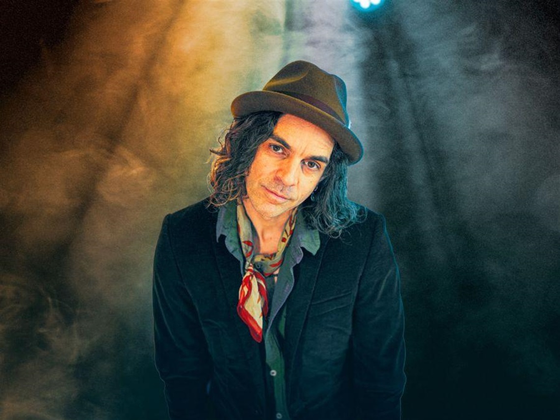 Nathan Cavaleri wearing dark jacket, blue collared shirt and brown hat under a rainbow spotlight against a dark hazy background