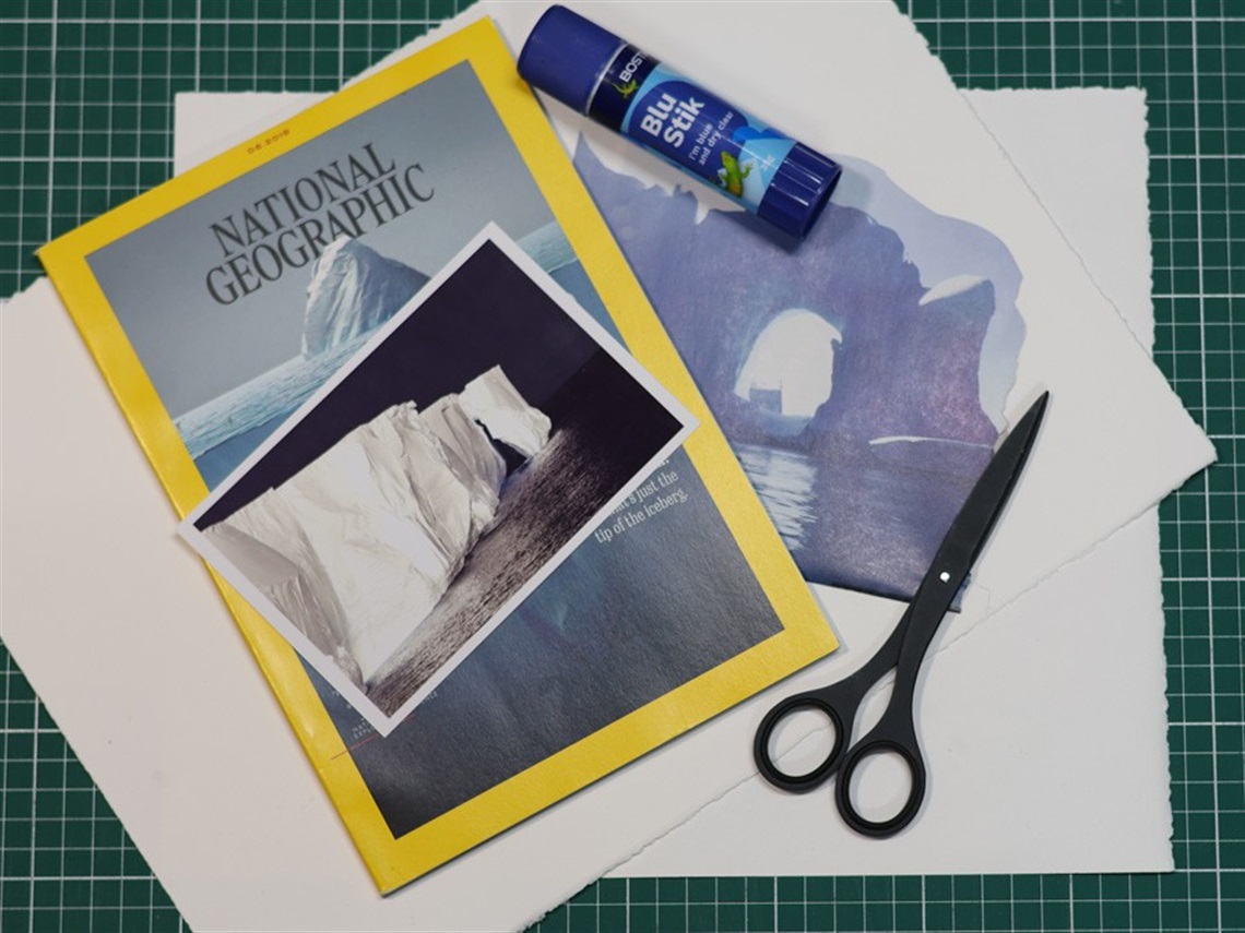 A green cutting board, blank pages of white paper, scissors, a glue stick as well as the cover of a National Geographic magazine (showing an iceberg), a postcard sized photograph of an iceberg and a page torn from an advertisement for a trip to Antarctica (featuring an iceberg). 