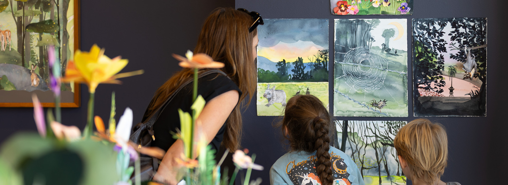 An adult and two children in a gallery, looking at watercolour paintings